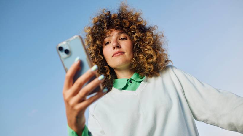 Person with curly blonde hair wearing white jacket and green collar holds up smartphone against bright blue sky background