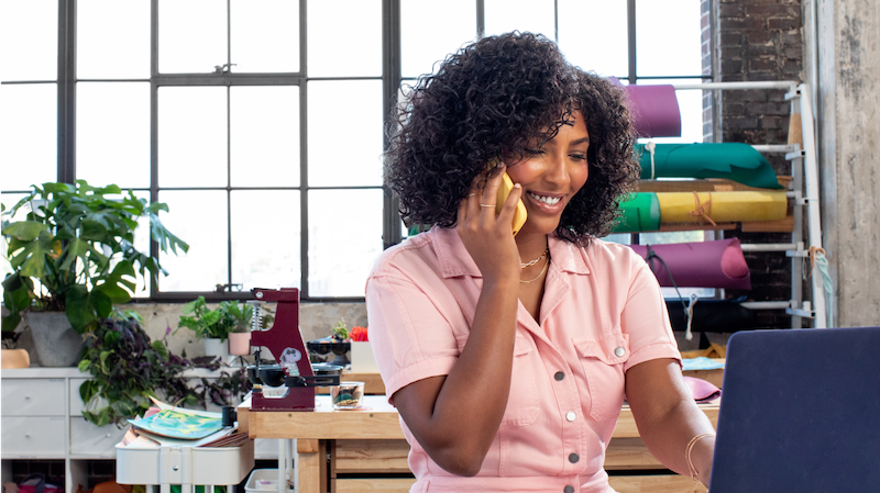 A woman in an office, speaking on her cell phone while she works on her laptop.